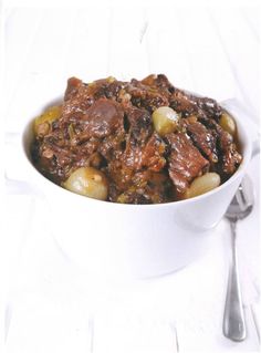 a white bowl filled with meat and potatoes on top of a table next to a fork