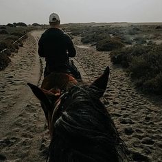 a man riding on the back of a brown horse down a dirt road next to a field