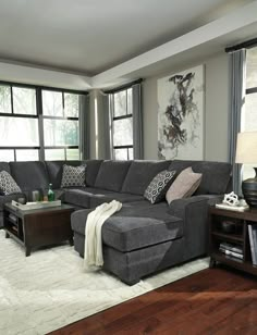 a living room with gray couches and pillows on top of the sofa, in front of large windows