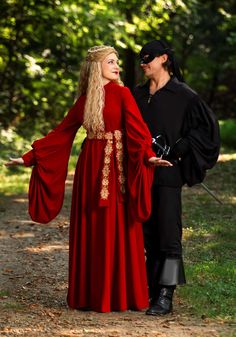 a man and woman dressed in medieval costumes walking down a path together, with trees in the background