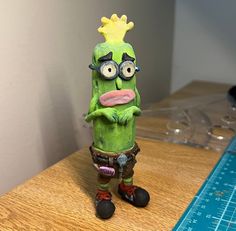 a green toy with googly eyes and a crown on it's head sitting on a table