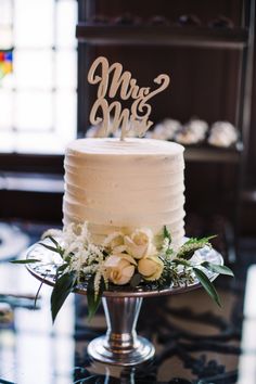 a wedding cake with white flowers and mr and mrs topper on a table in front of a stained glass window