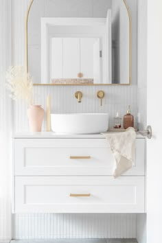 a white bathroom with gold accents and a round mirror above the sink, on top of a dresser