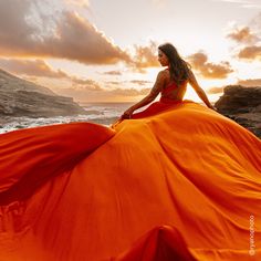 a woman in an orange dress is sitting on the rocks by the ocean at sunset