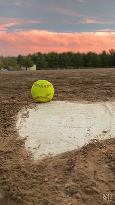 a yellow ball sitting on top of a dirt field next to a baseball field with trees in the background