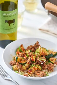 a white bowl filled with meat and vegetables next to a bottle of wine on a table