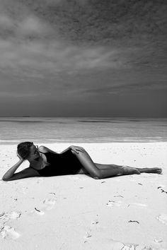 a woman laying on top of a sandy beach next to the ocean in black and white