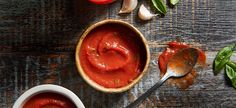 two bowls filled with tomato sauce on top of a wooden table next to garlic and tomatoes