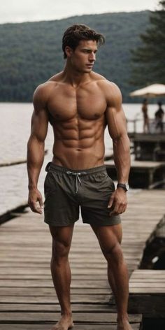 a shirtless man standing on a dock by the water with his hands in his pockets