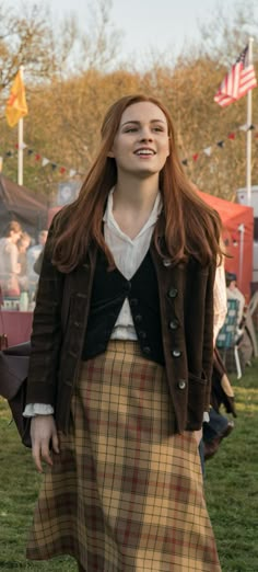 a woman with long red hair wearing a brown jacket and plaid skirt standing in the grass