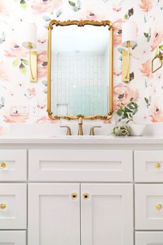 a bathroom with floral wallpaper and gold framed mirror above the sink, along with white cabinets