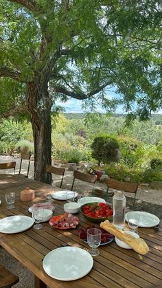 a wooden table with plates and glasses on it in front of a tree filled field