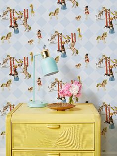 a yellow dresser sitting next to a blue lamp on top of a wooden table in front of a wallpapered background