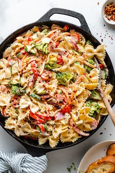 a skillet filled with pasta and vegetables next to garlic bread on a white surface