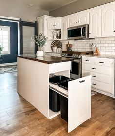 a kitchen with white cabinets and wooden floors has an open drawer in the middle that holds two black trash cans