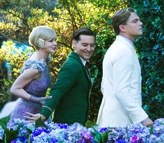 two men and a woman standing next to each other in front of some bushes with trees behind them