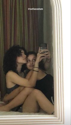 two women are sitting on the bed taking a selfie