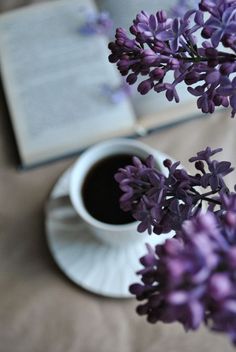 a cup of coffee next to an open book and some purple flowers on a table