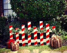 a red and white striped fence decorated with christmas presents in front of a house, surrounded by greenery