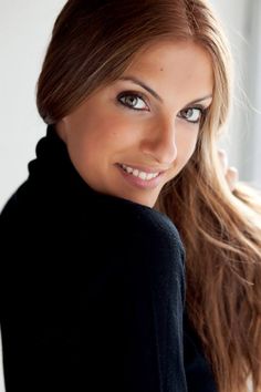 a woman with long brown hair and blue eyes smiles at the camera while leaning against a wall