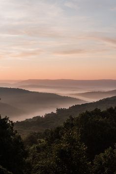 the sun is setting over some trees and hills with mist in the valley behind it