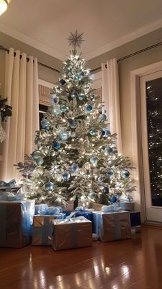 a white christmas tree with blue and silver presents under it in front of a window