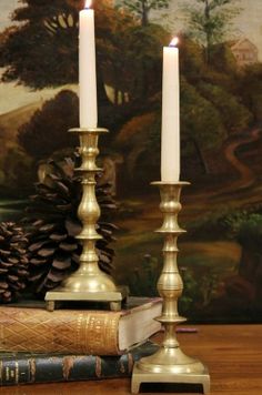 two candles sitting on top of an old book next to some pine cones and a painting