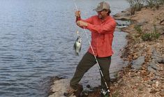 a man holding a fish while standing next to a body of water with a fishing pole