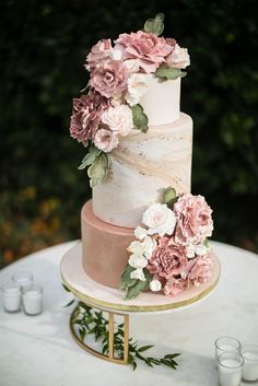 a three tiered cake with pink flowers and greenery sits on a gold stand