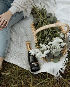 a woman sitting on the ground next to a basket with flowers and a bottle of champagne