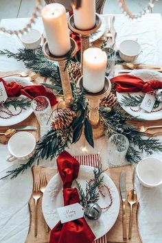 a christmas table setting with pine cones, candles and red ribbon on the place settings