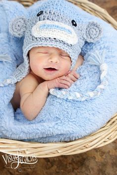 a baby wearing a crocheted hat laying in a basket with a stuffed animal