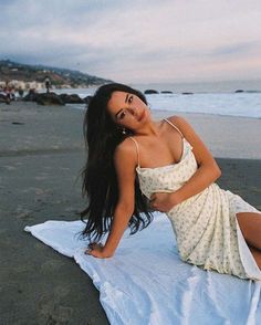 a beautiful woman laying on top of a white towel near the ocean with her legs crossed