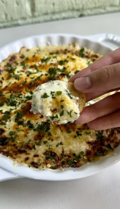 a hand holding a piece of bread with cheese and herbs on it in a white bowl