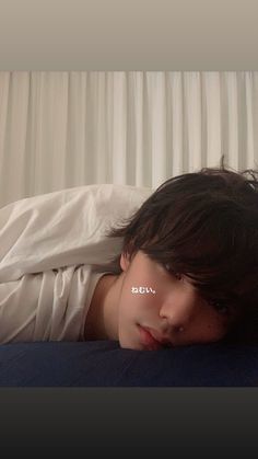 a young man laying on top of a bed next to a white curtained window