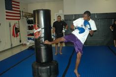 a young man kicking a punching bag in a gym with two other men standing behind him