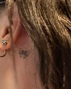 a woman with a small bow tattoo on her left ear and behind the ear is a pair of gold hoop earrings