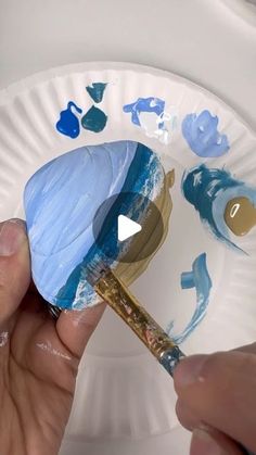 someone is painting on a paper plate with blue and gold paint, while another hand holds a brush