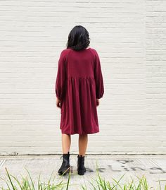 a woman standing in front of a white brick wall wearing a maroon dress and black boots