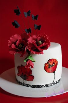 a white cake decorated with red flowers and black leaves on a plate against a red background