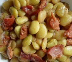 a white bowl filled with beans and bacon on top of a wooden table next to a fork