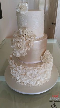 a three tiered wedding cake with white flowers on the top and bottom, sitting on a glass table