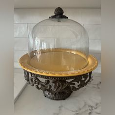 a cake stand with a glass dome on top and gold trim around the base, sitting on a marble countertop