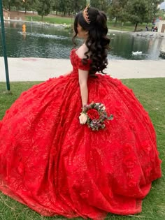 a woman in a red ball gown sitting on the grass near a body of water