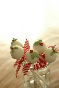 four white cake pops in a glass vase with red and green bows on them, sitting on a table