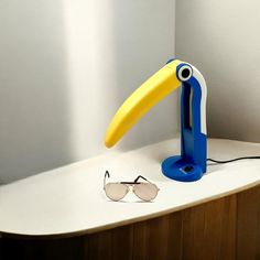 a blue and yellow bird shaped lamp next to sunglasses on a white counter with a pair of glasses