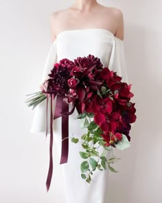 a woman wearing a white dress holding a bouquet of red flowers and greenery in her hands