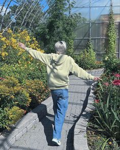 an older woman walking down a sidewalk in front of some plants and flowers with her arms outstretched