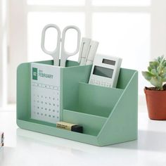 a desk with a calendar, pen and calculator on it next to a potted plant