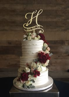 a white wedding cake with red and white flowers on the top tier is displayed against a wooden wall